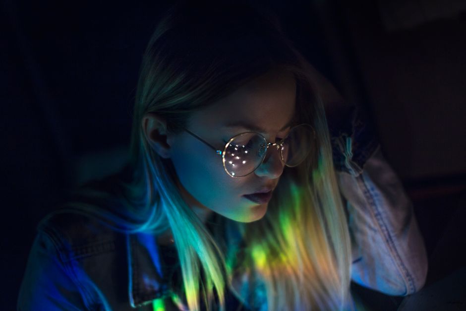 A woman in glasses with colorful reflections on her face in a dimly lit setting.