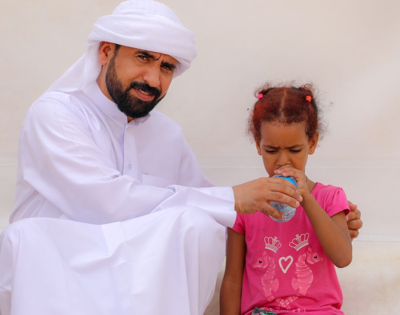 A man in traditional attire helps a young girl drink water, showcasing kindness and cultural attire.