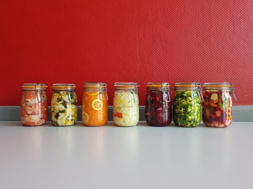 A row of vibrant, multicolored mason jars filled with assorted fermented vegetables against a red wall.