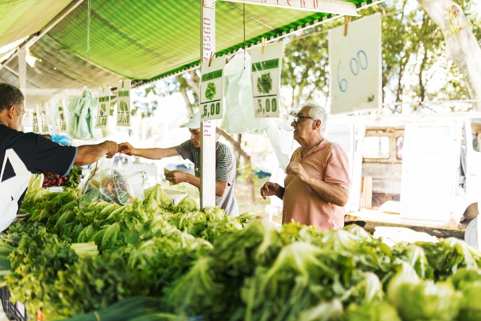 Customers purchasing fresh vegetables at an outdoor farmers market under green canopy.
