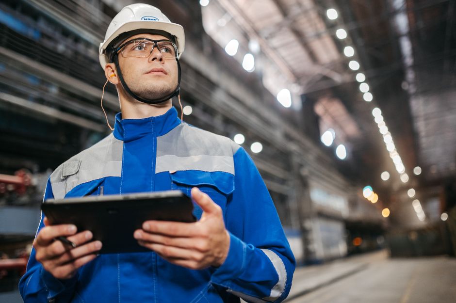 Factory worker in safety gear using a tablet in a well-lit industrial setting.