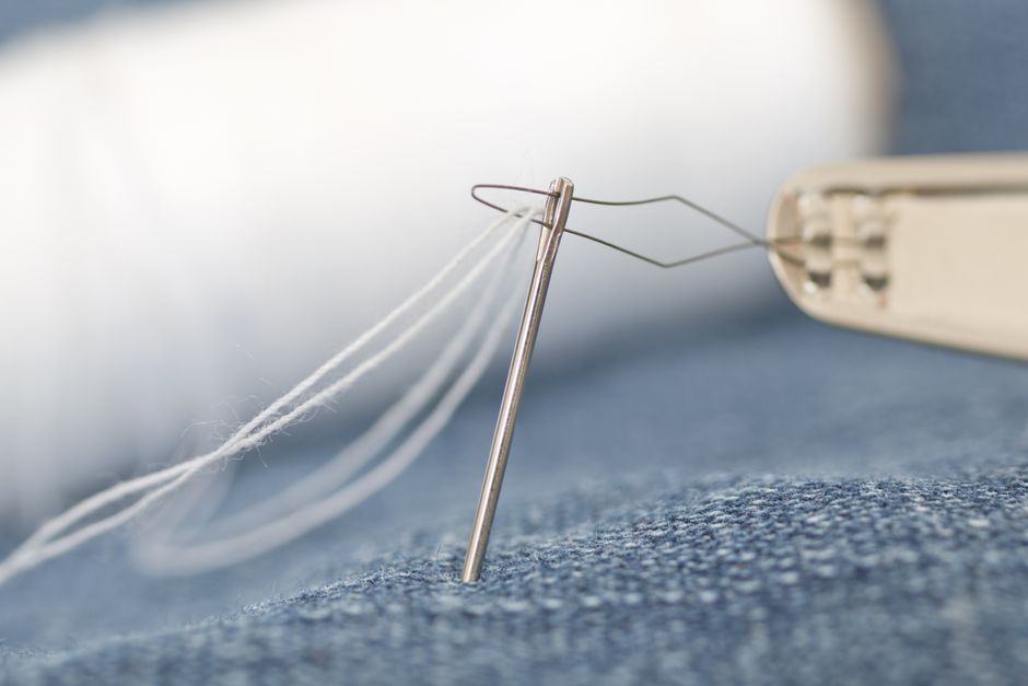 Detailed macro shot of a sewing needle threading white thread through denim fabric.