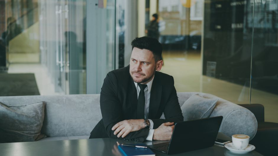 A businessman in a suit sits thoughtfully in an office lounge with a laptop.
