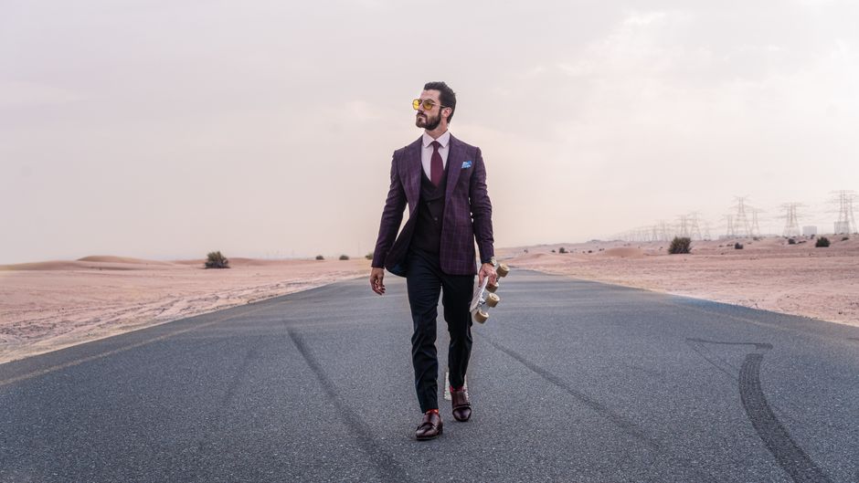 Man in formal wear walking on deserted road holding a skateboard, showcasing stylish fashion in a desert setting.