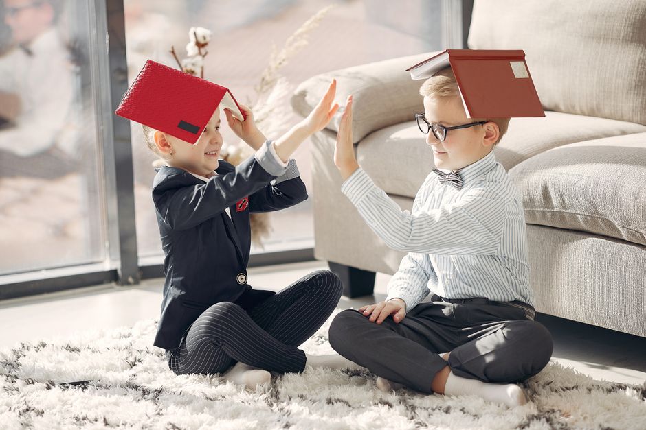 Two children playfully balance books on their heads while high-fiving indoors, capturing joy and creativity.