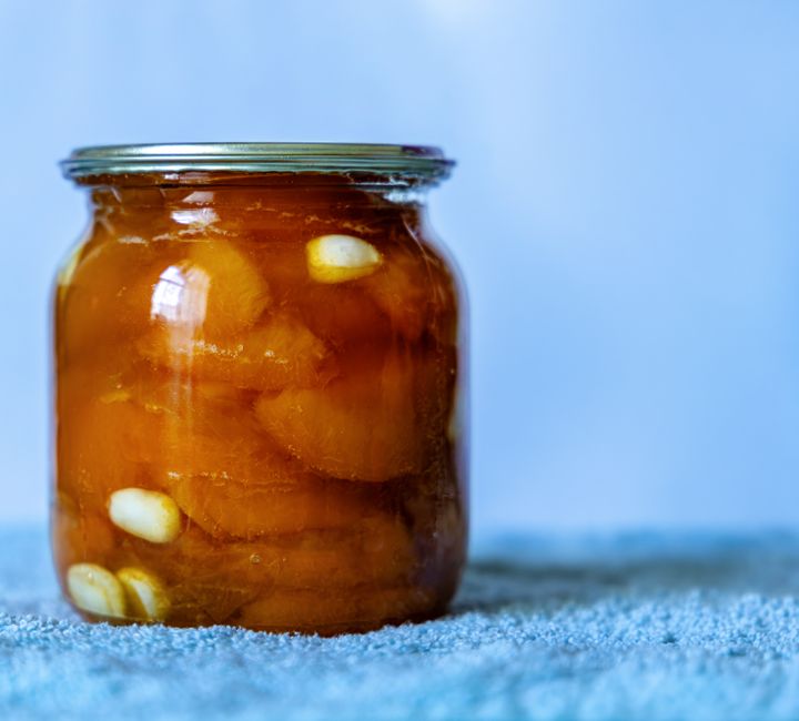 A close-up of a jar filled with preserved garlic and peaches, set against a soft blue background.