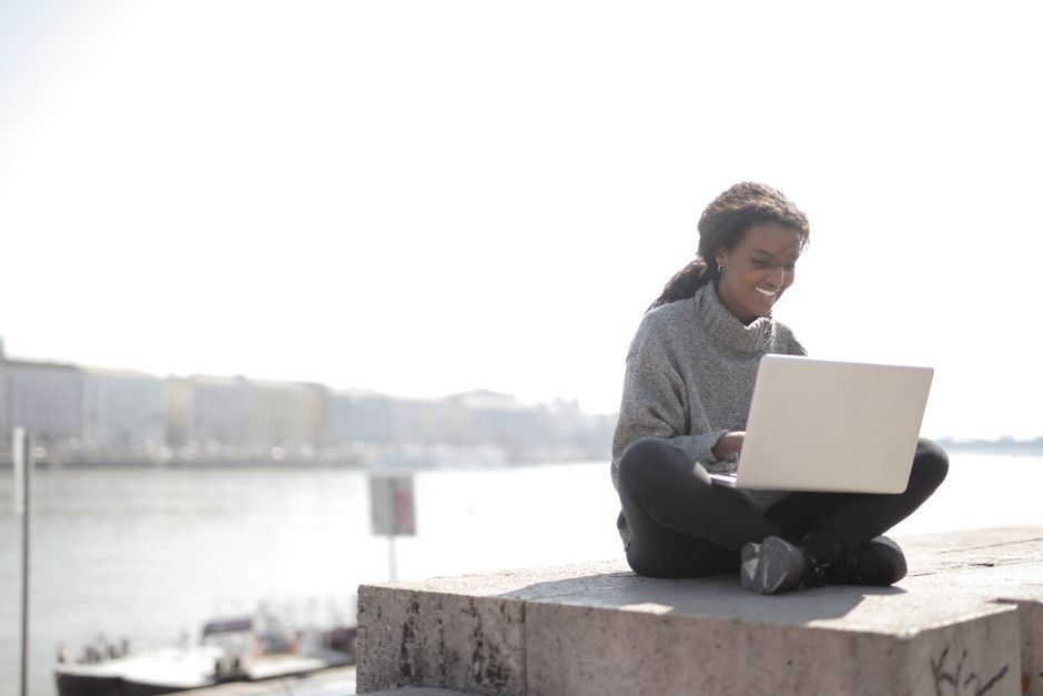 Smiling woman freelancing on a laptop by a riverside cityscape, representing remote work leisure.
