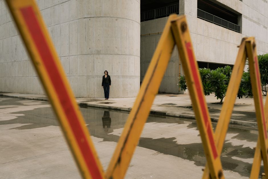 A lone person stands against a modern concrete building, viewed through a barrier.