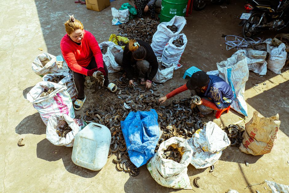A diverse group sorts through scrap metal in an outdoor market setting, surrounded by bags of recyclables.
