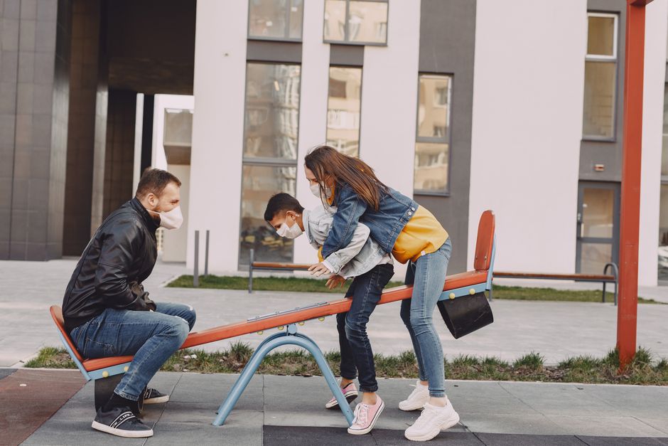 Side view of positive family in trendy casual and medical mask clothes resting near modern building while swinging together on seesaw on urban playground during daytime