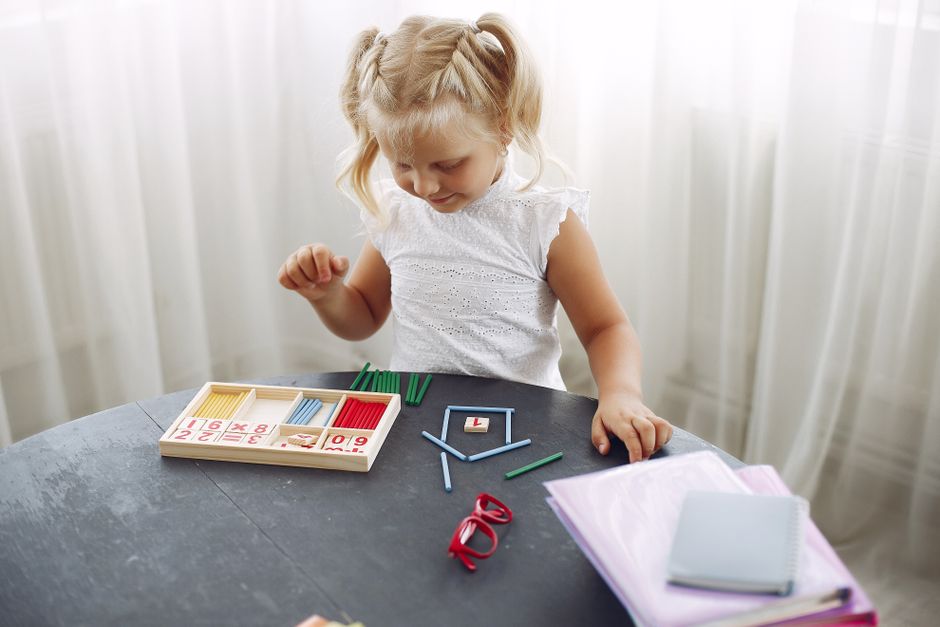 Blond little girl standing while playing with puzzle in living room at home and counting sticks different colors during weekend