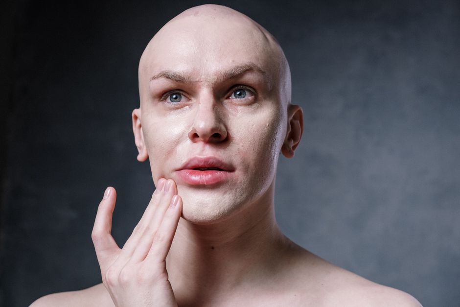 A bald man applies makeup in a studio setting, focusing on skincare and beauty routines.
