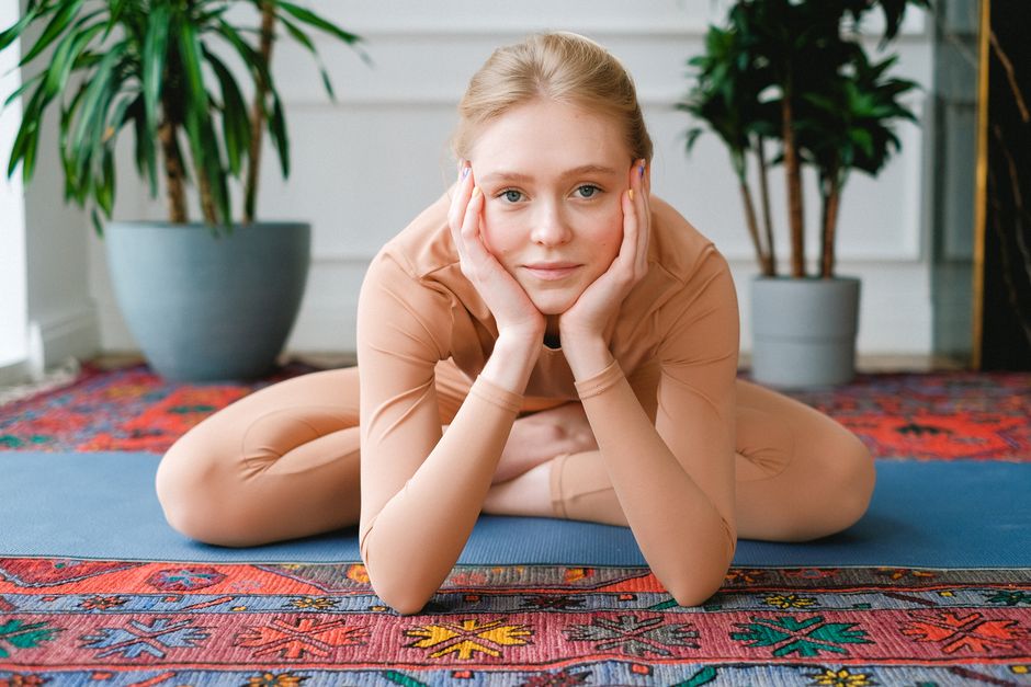 Full length positive female in beige sportswear sitting in Lotus Pose on yoga mat and leaning on hands while looking at camera at home
