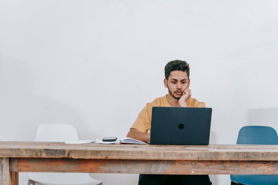 Focused young Hispanic male student watching online lecture on laptop while sitting at wooden table with notebooks during remote lesson