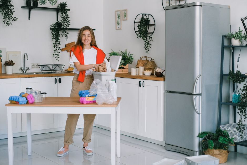 Smiling female in casual clothes standing near table and looking at camera while sorting rubbish in light cozy kitchen at home