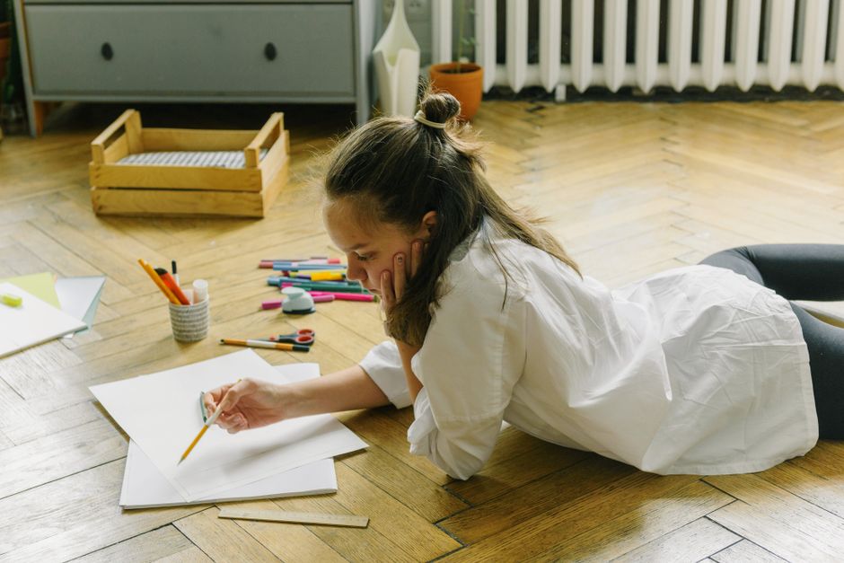 Teen girl draws on floor surrounded by art supplies, focusing on creative expression.