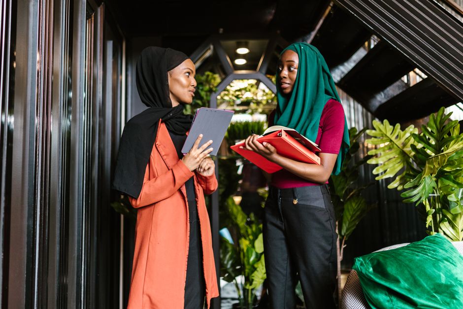 Two women in hijabs discuss business in a modern indoor setting.
