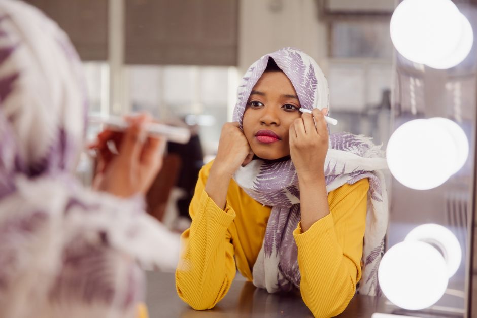 Portrait of a young woman in a hijab applying makeup at a dressing table with lights.