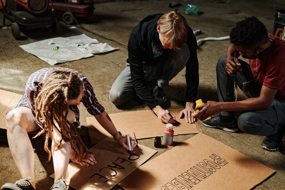 Three adults making protest signs with cardboard and markers indoors.