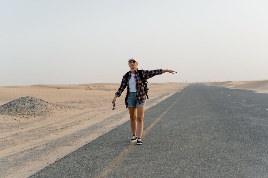 A young woman with a backpack hitchhiking on an open desert road under clear skies.