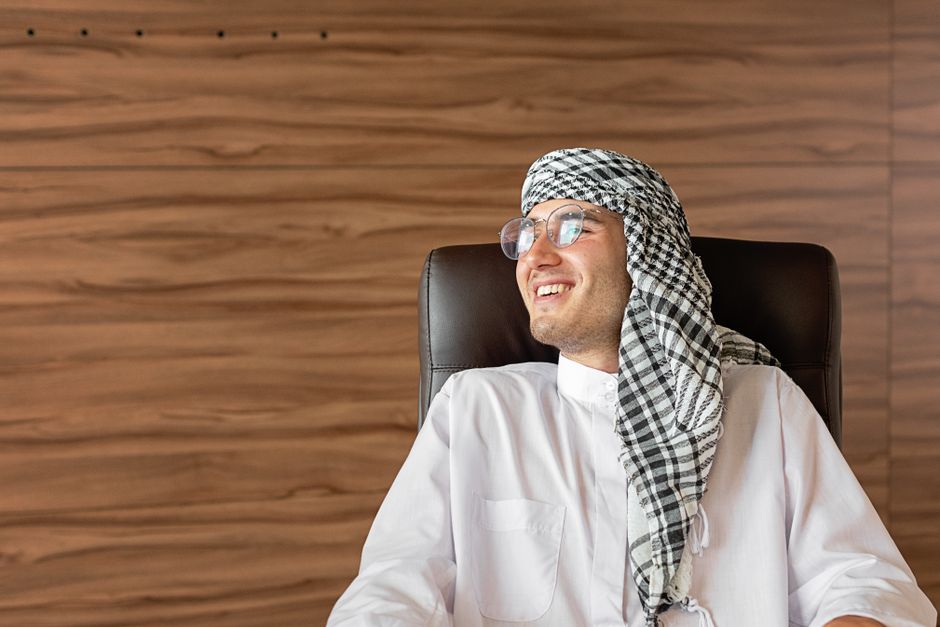 Cheerful man with headscarf sitting in an office chair indoors.