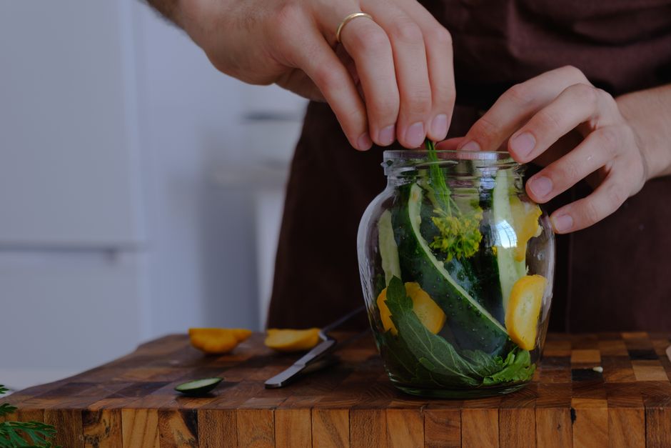 Hands organizing fresh vegetables for pickling in a glass jar on a wooden countertop.