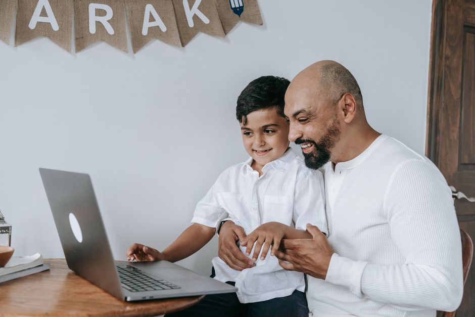 A father and son smiling while using a laptop, creating a moment of joy and bonding indoors.