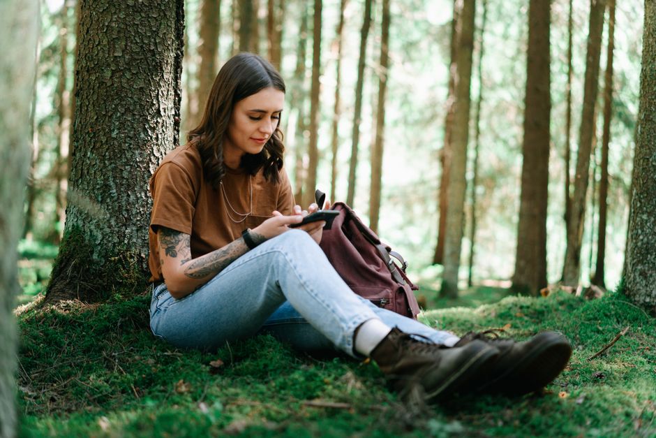 Young woman sitting in a forest using a smartphone, surrounded by nature. Relaxation and adventure.